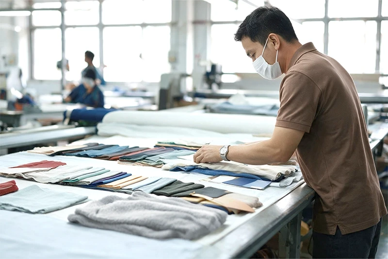 A masked male textile professional carefully examines various colored fabric swatches of polyester fleece, circular knit, and poly boa on a long cutting table inside a garment factory, with other workers visible in the background.